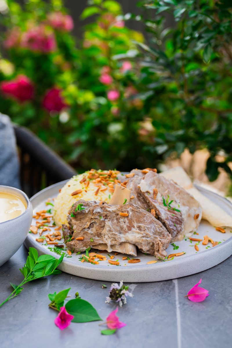 A lamb and rice dish garnished with fried almonds and herbs on a grey plate, outdoors with flowering bushes behind