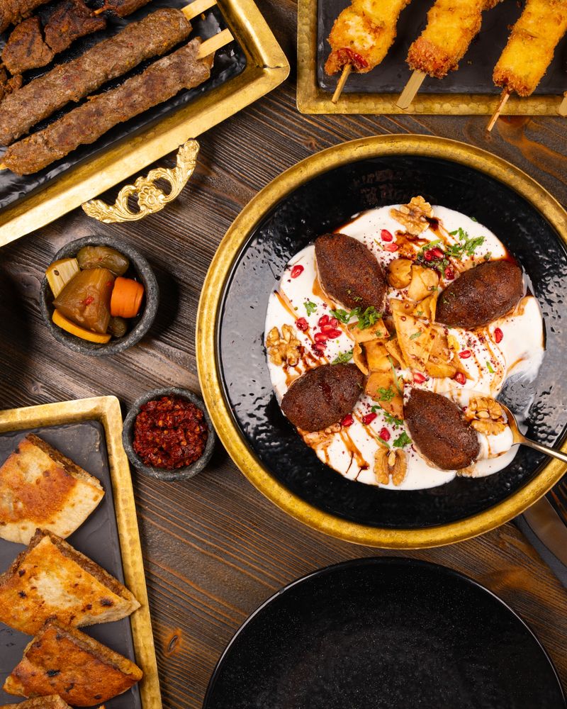 Overhead view of a Middle Eastern table: four dark fried croquettes in white yoghurt with fried bread and pomegranate seeds, grilled skewers, pickles and flatbreads on gold trays