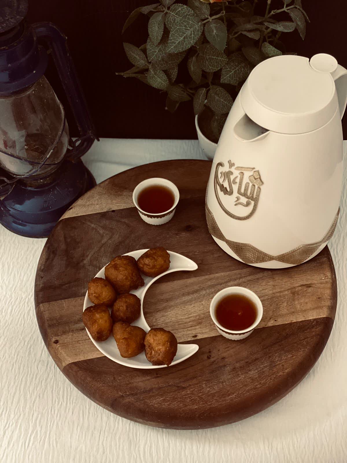 Luqaimat on a crescent-shaped white plate beside two small cups of Arabic coffee and a white coffee flask on a wooden board