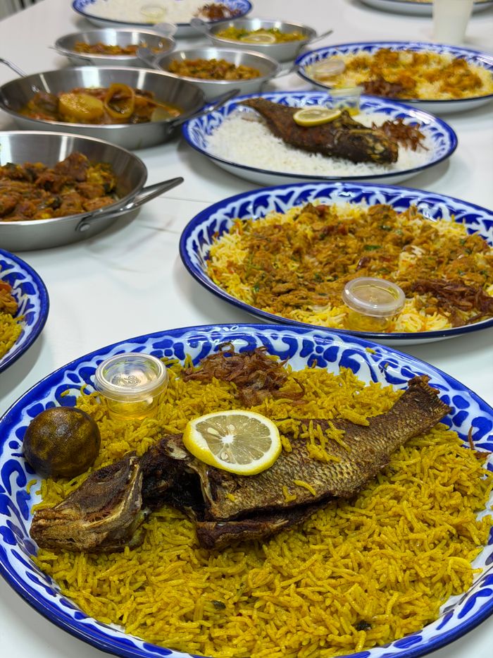 Fried fish on saffron-yellow rice with a lemon slice and a whole dried lime on a blue-and-white platter, with other dishes behind