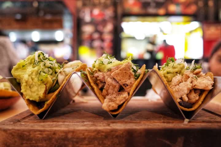 Three tacos with chicken, guacamole and chopped onion standing in metal holders on a wooden board