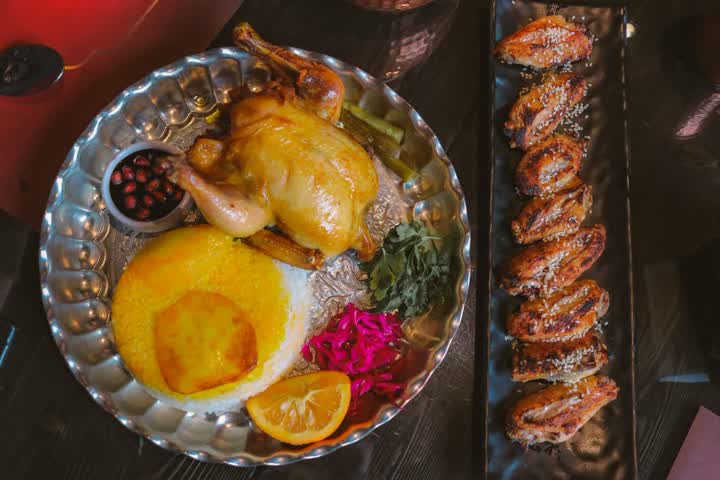 A whole roast chicken on a silver platter with saffron rice, a pomegranate dip and herbs, beside a tray of grilled chicken wings, seen from above