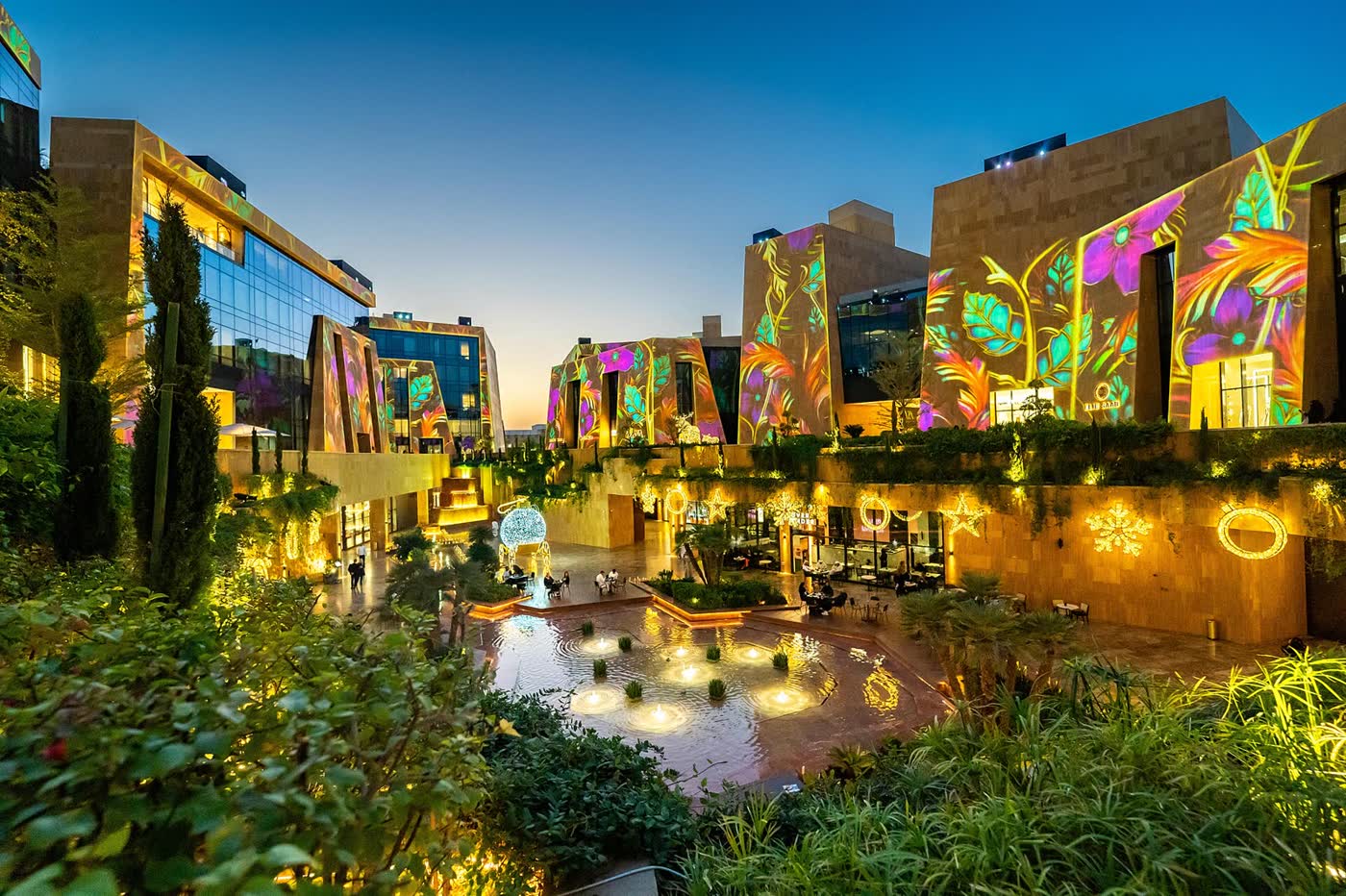 VIA Riyadh at dusk, with sculpted stone buildings lit by projected floral patterns around a garden courtyard