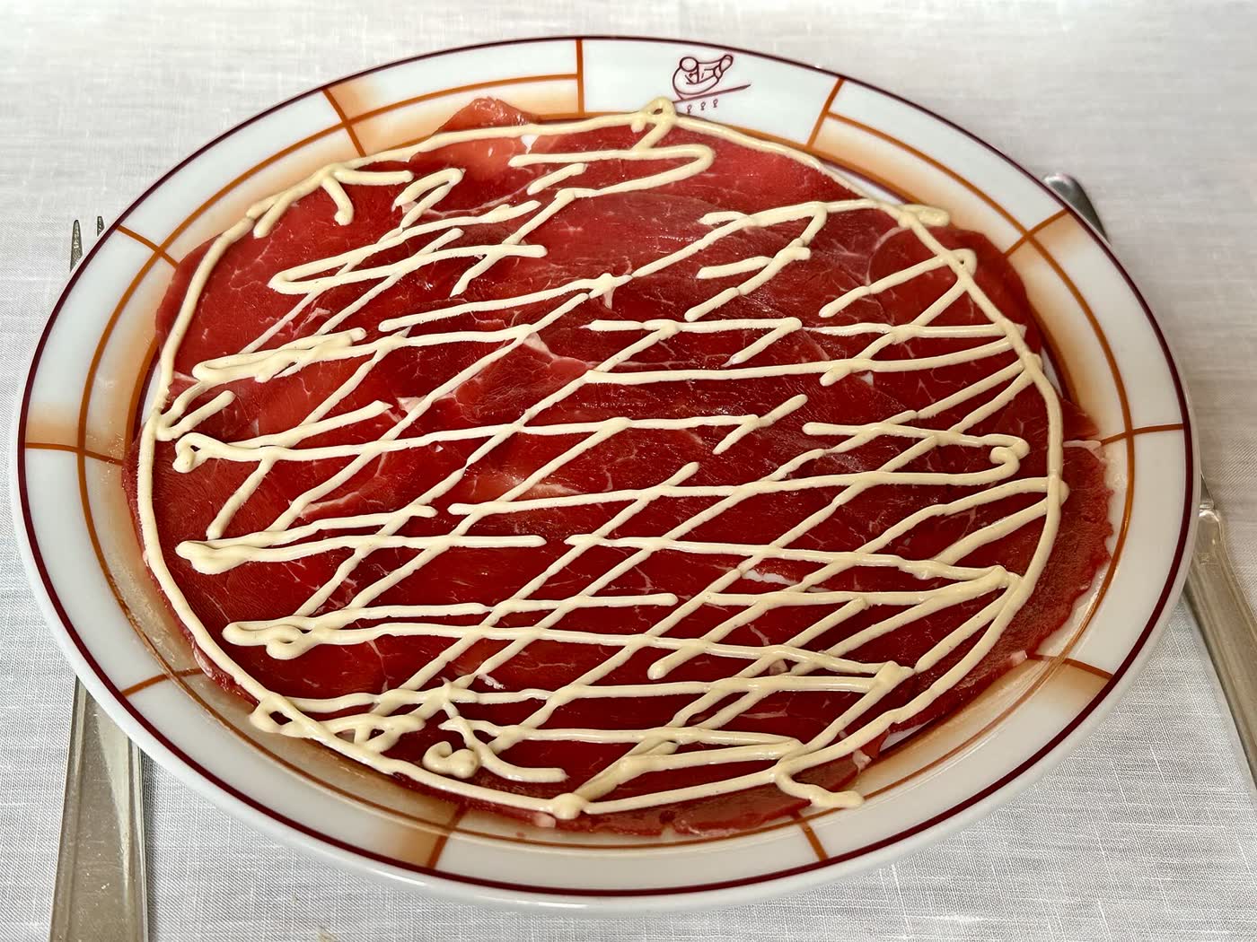 Carpaccio of thinly sliced beef on a white plate, drizzled with a lattice of pale sauce