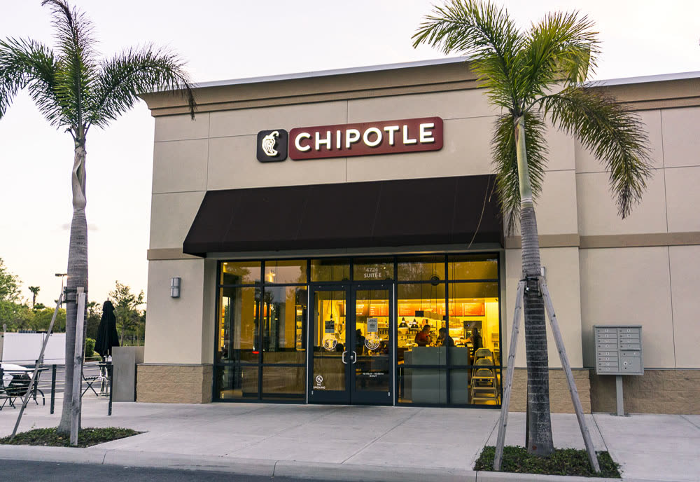 A Chipotle restaurant with a dark awning and palm trees outside, photographed at dusk