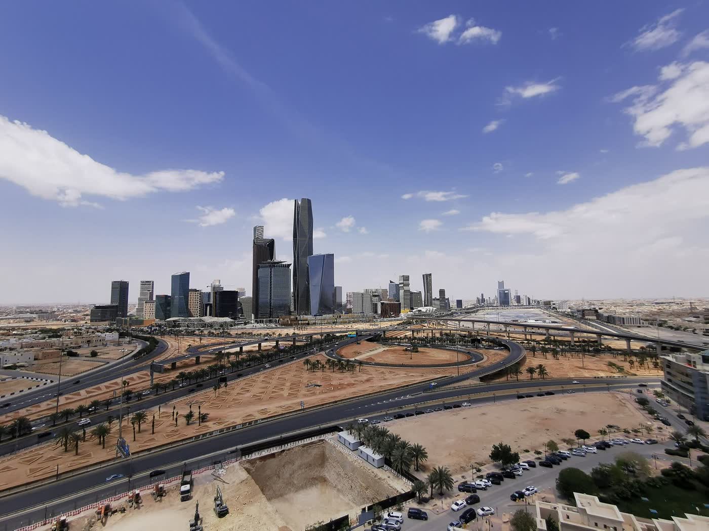 The skyline of the King Abdullah Financial District in Riyadh under a blue sky with scattered clouds, seen above a road interchange