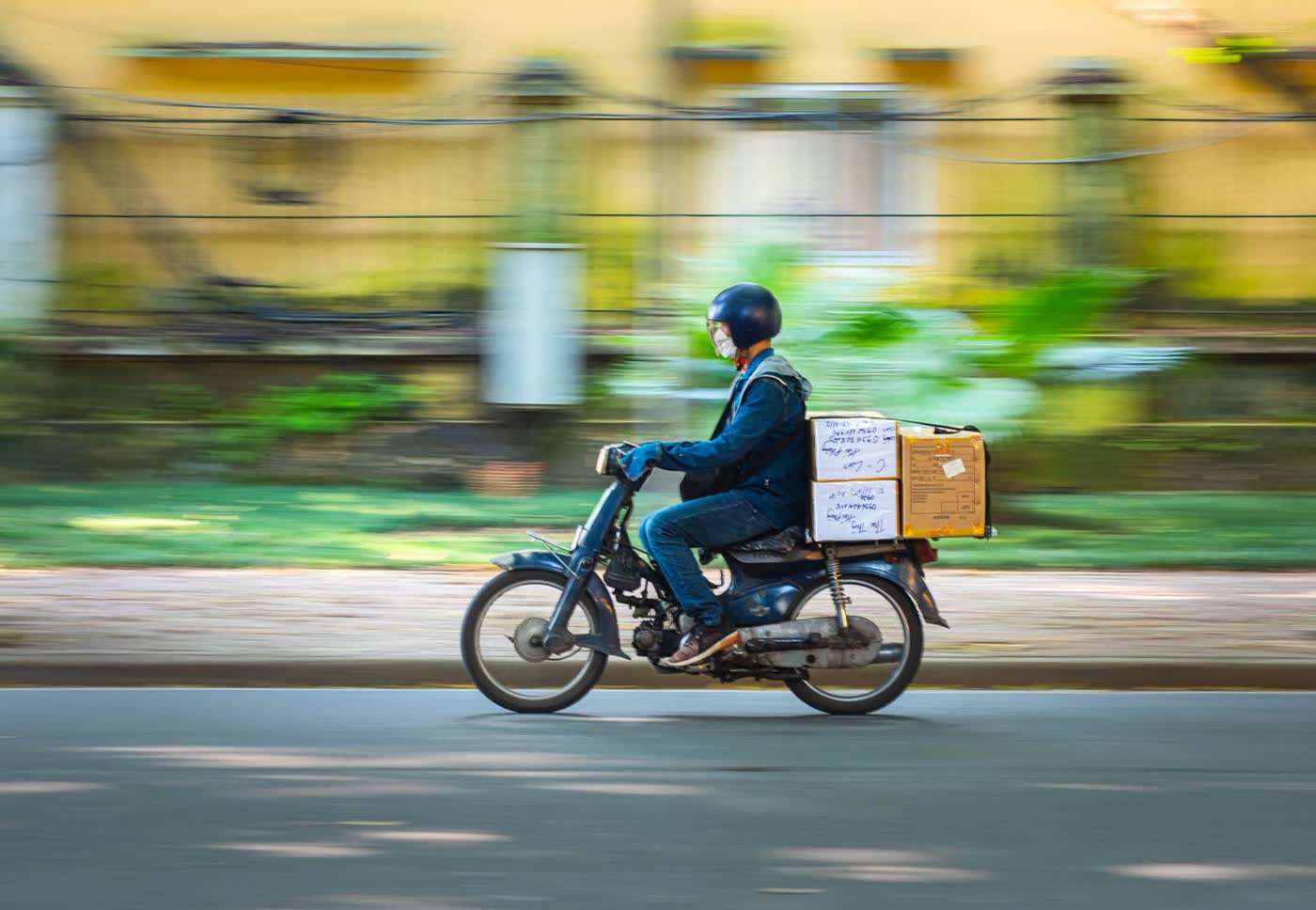 A courier in a blue helmet and jacket riding a small motorbike carrying two cardboard boxes, with a blurred yellow wall behind