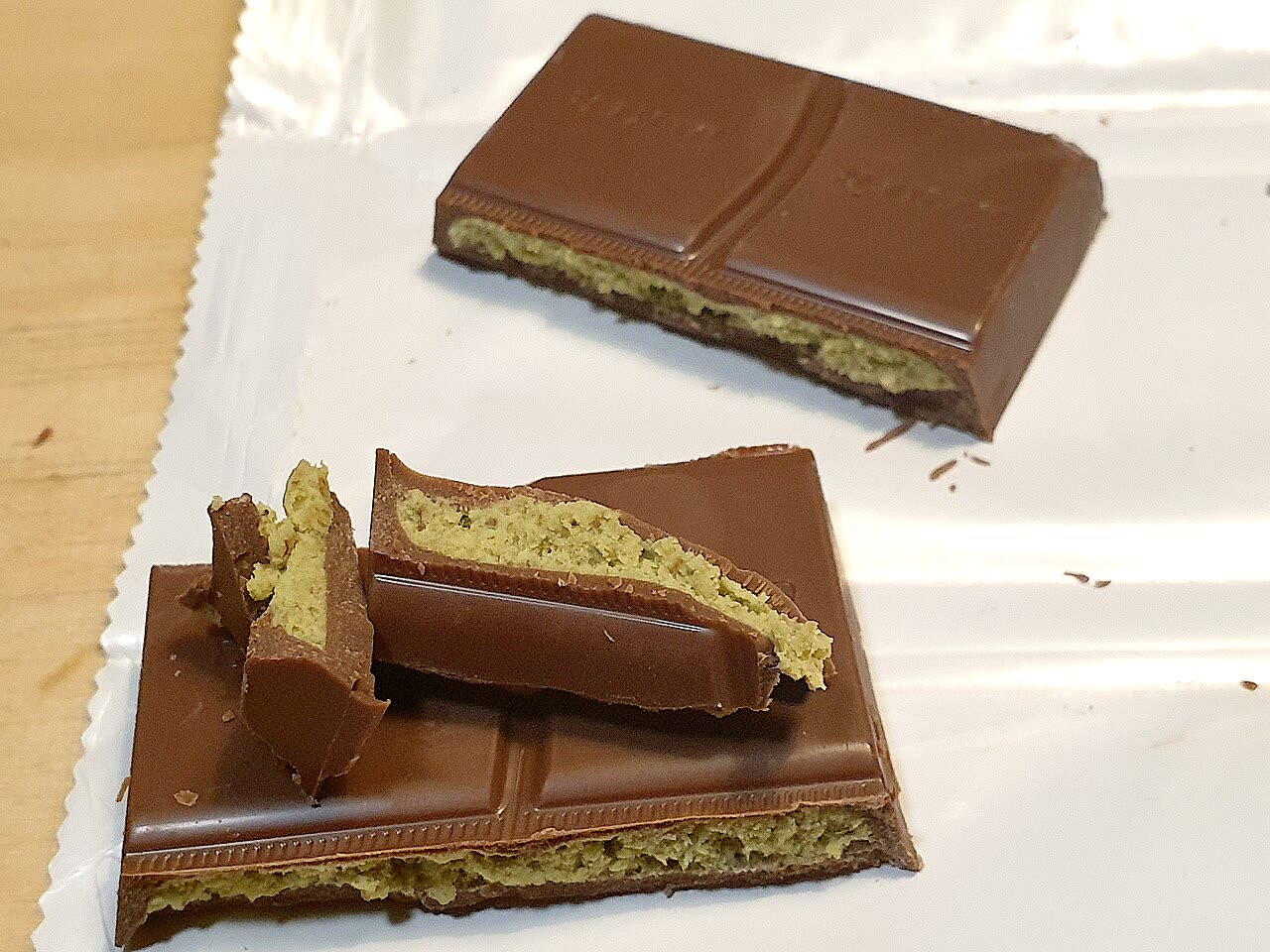 A milk chocolate bar broken into pieces on foil, showing a thin layer of green pistachio filling