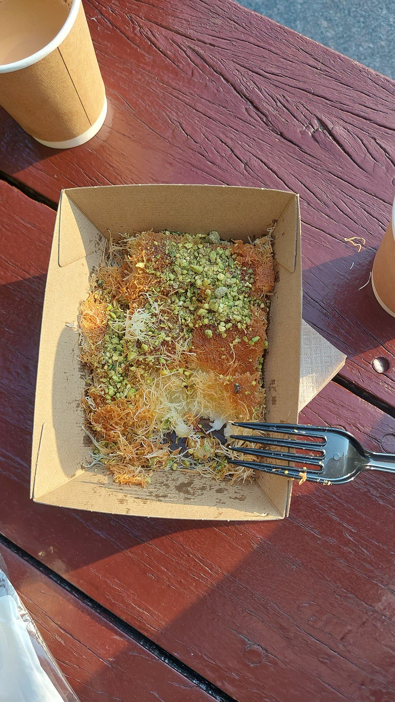 A cardboard box of kunafa topped with crushed green pistachios, with a black plastic fork resting in it