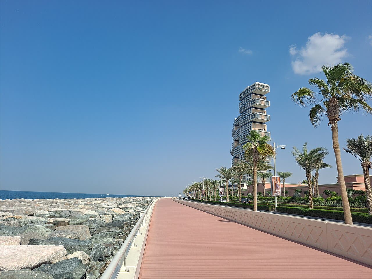 A boardwalk and palm-lined road along the sea on the Palm Jumeirah, with the stacked tower of Atlantis The Royal in the distance