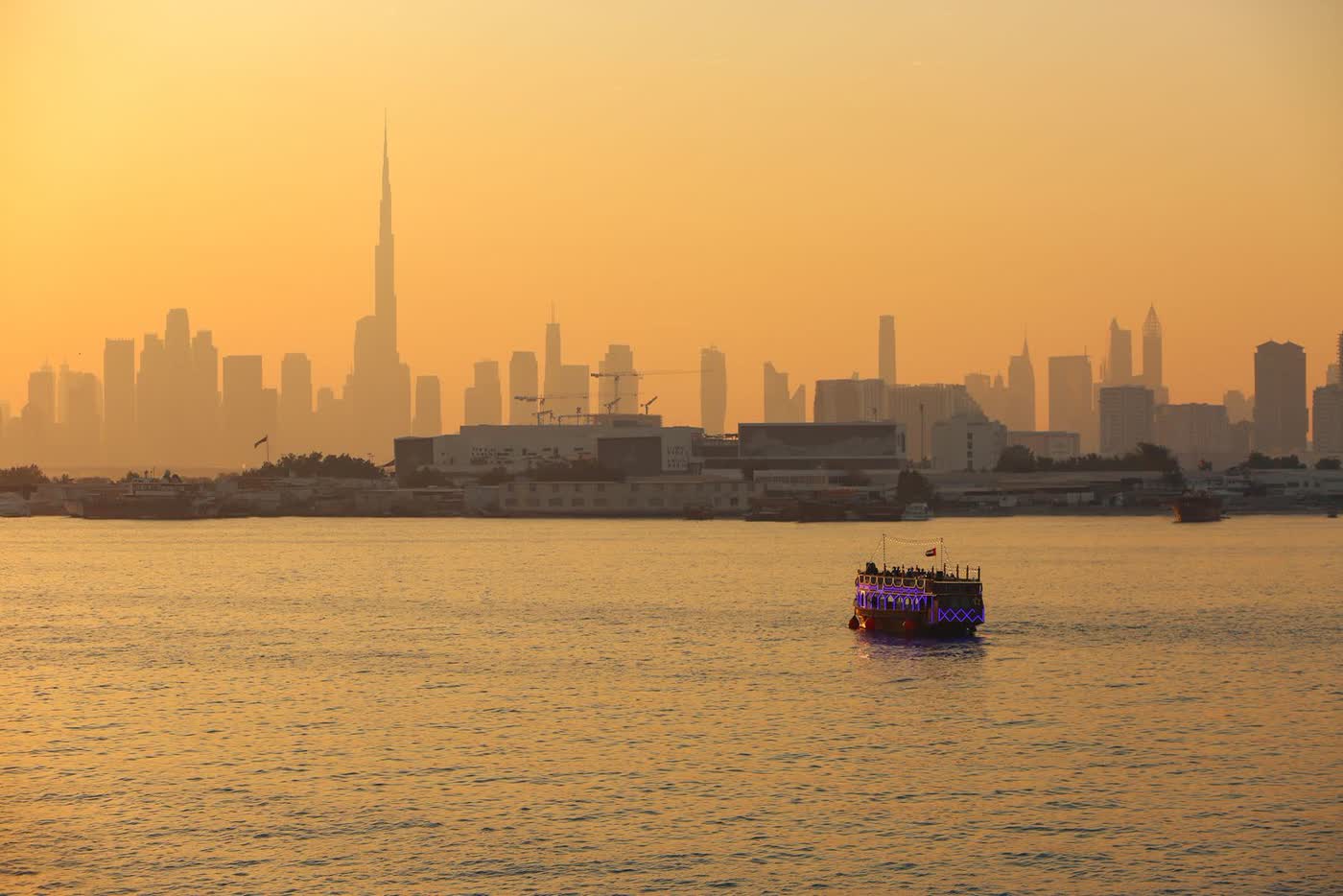 Dubai’s skyline with the Burj Khalifa in golden evening haze, seen across the water, with a lit dhow in the foreground