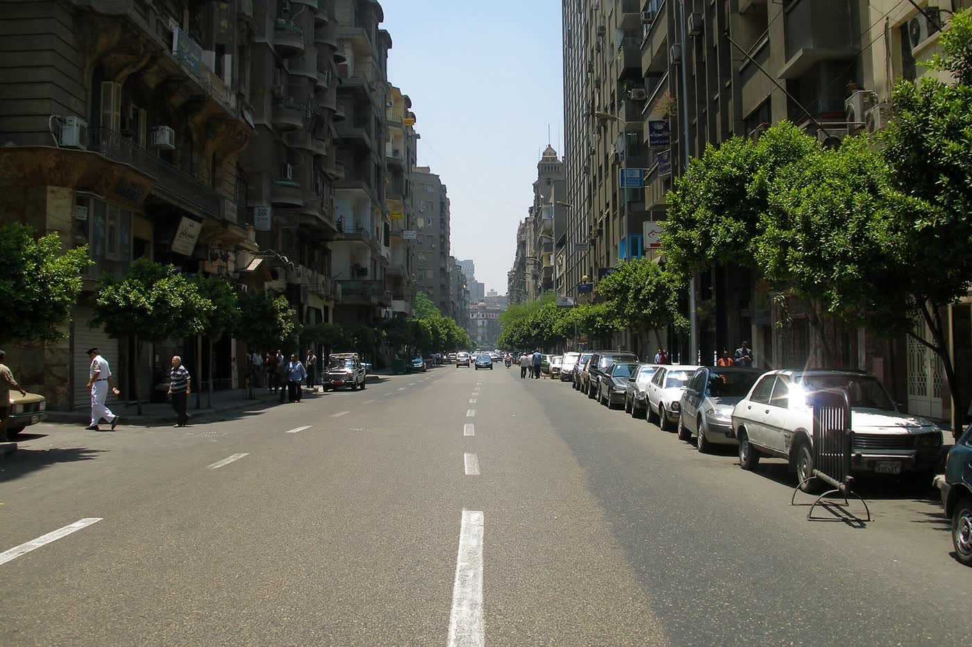 A long, tree-lined city street in Cairo with tall apartment blocks on both sides and parked cars along the kerb