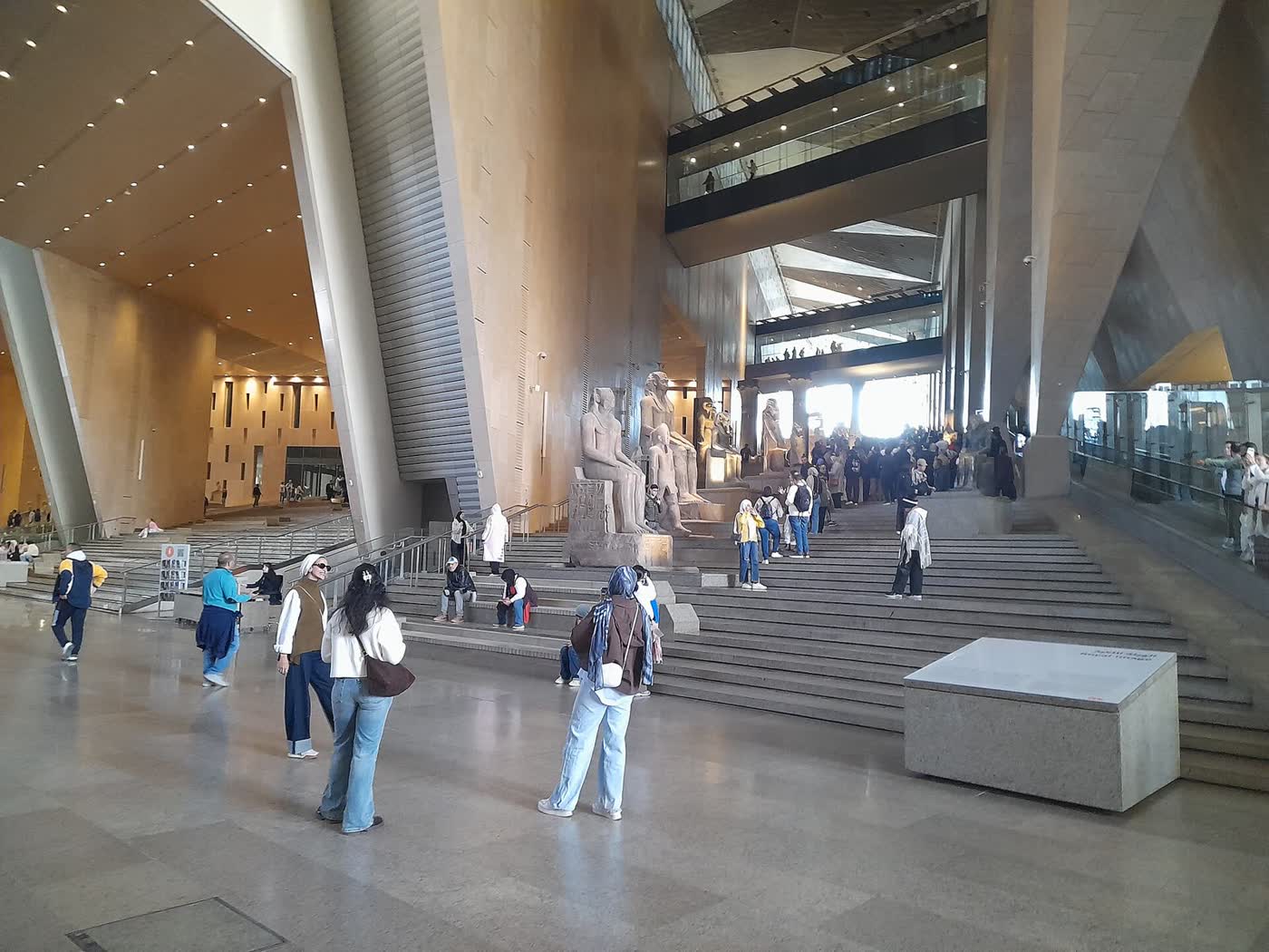 Visitors on the stone steps of a vast museum hall, with seated royal statues and walkways high above