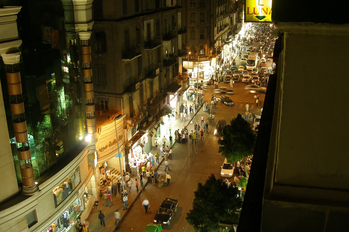 A busy Cairo street at night seen from above, with shop fronts, crowds on the pavement and traffic stretching into the distance