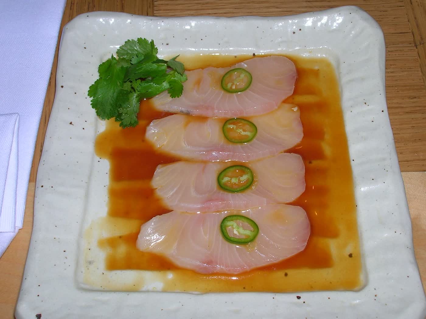 Four slices of pale yellowtail sashimi, each topped with a round of green chilli, on a square white plate with a sprig of coriander