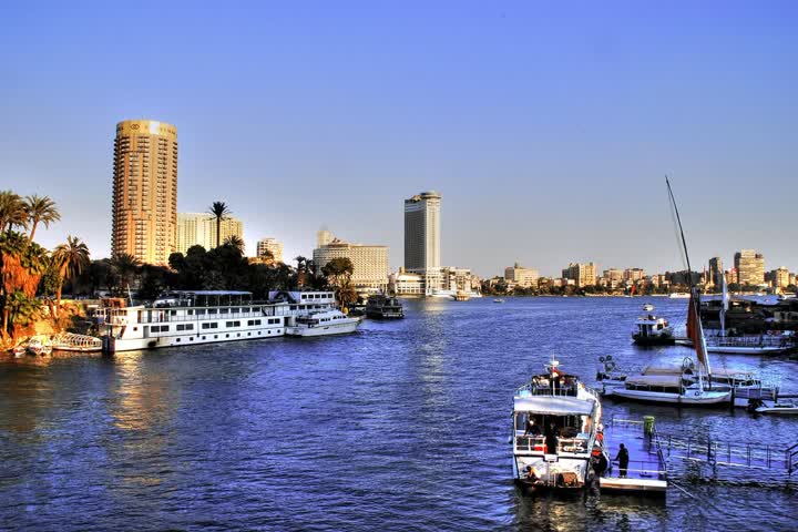 The Nile at Cairo in evening light, with boats moored along the banks, a tall golden tower and hotel blocks on Gezira Island