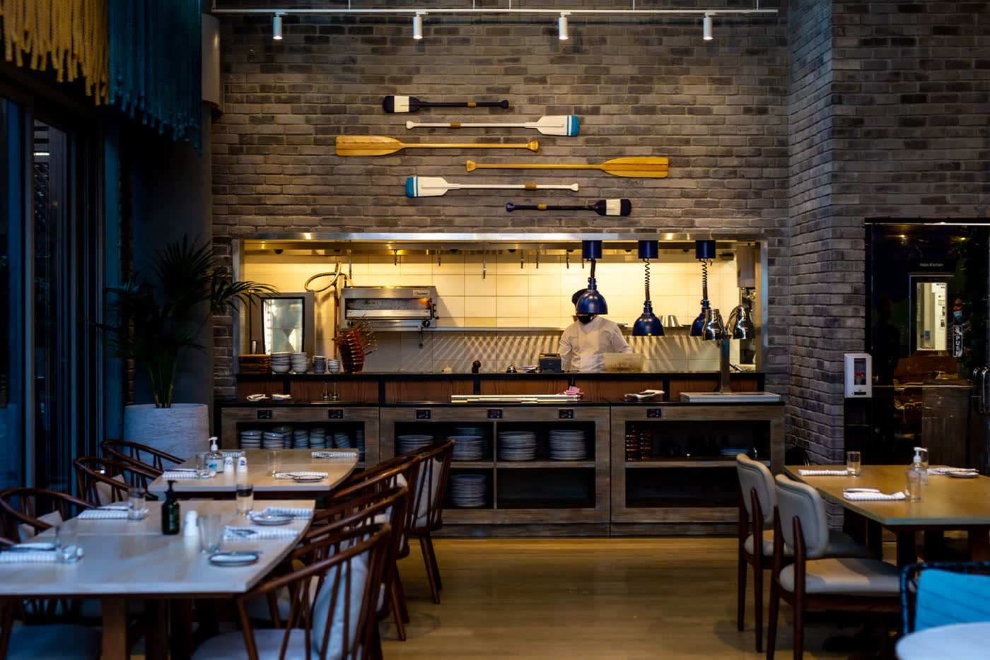 A restaurant dining room with laid tables and an open kitchen behind a counter, with a chef at work