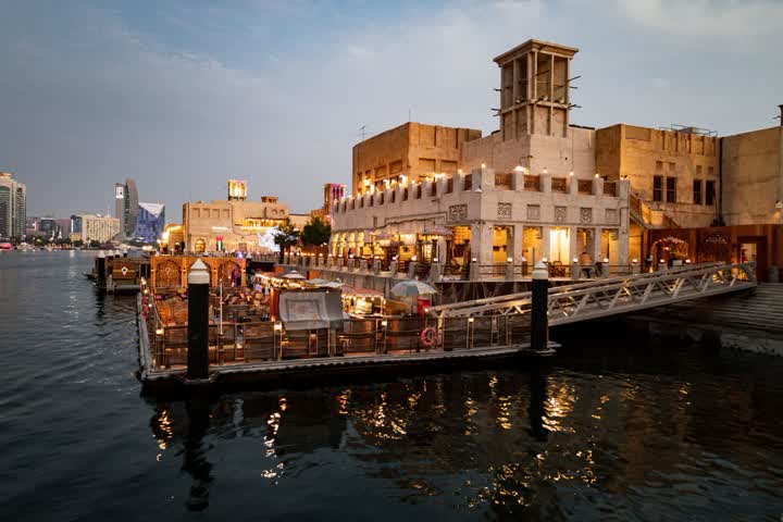 Floating restaurant terraces and an old wind-tower building beside the water in Dubai at dusk, lit by warm lights