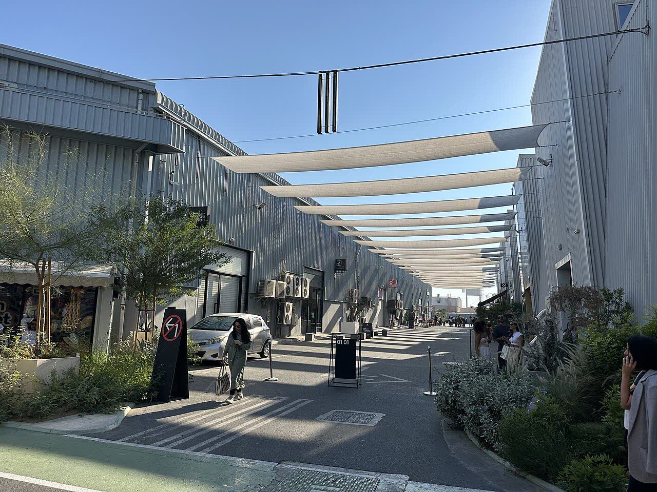 A pedestrian street between corrugated-metal warehouse buildings, shaded by long white sails strung overhead