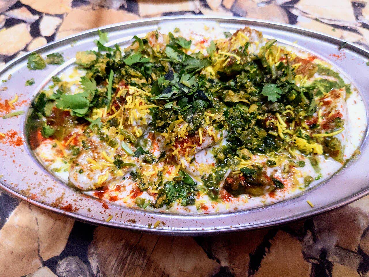 A steel plate of chaat covered in yoghurt, green chutney, red spice and fine yellow sev