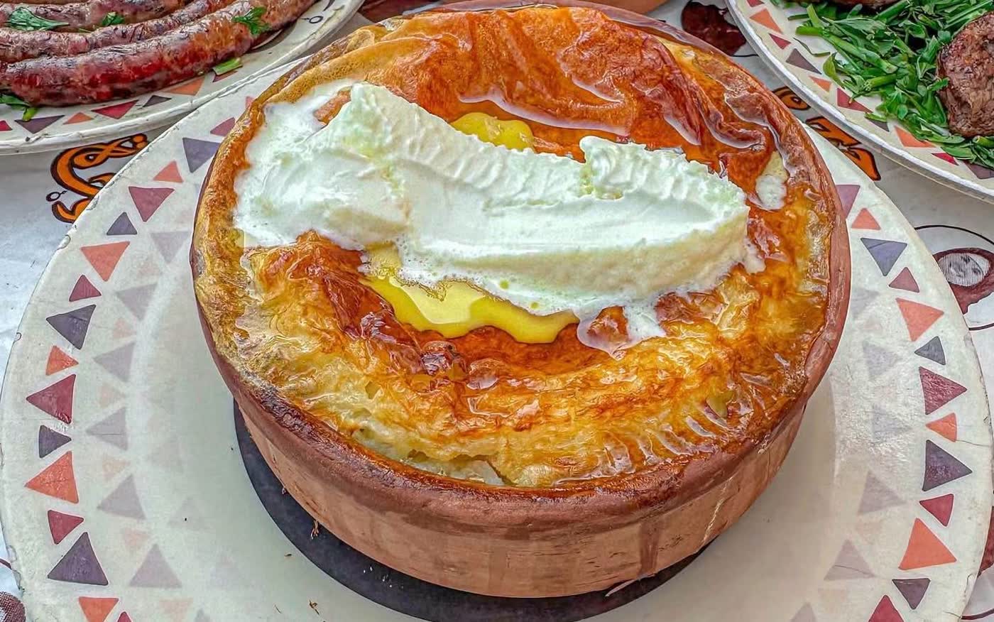 A clay pot of baked rice pudding with a deeply browned crust and a ribbon of cream on top, standing on a plate with a triangle-pattern rim