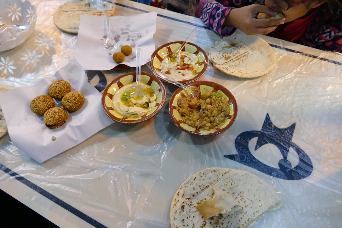 A table covered in clear plastic with bowls of hummus and other dips, sesame-crusted falafel on paper, and flatbread