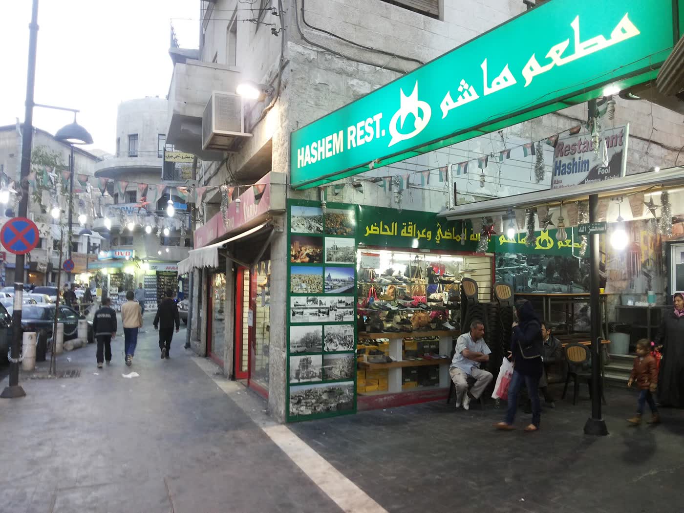 The green Hashem Restaurant sign in English and Arabic above a corner shopfront on a downtown Amman street, with pedestrians and strings of Jordanian flags