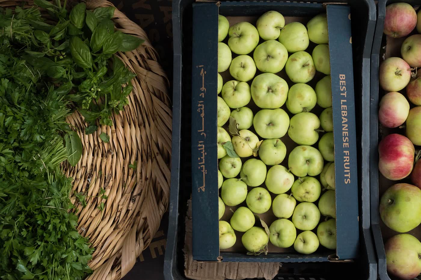 A crate of green apples labelled Best Lebanese Fruits in Arabic and English, beside a basket of fresh parsley and basil