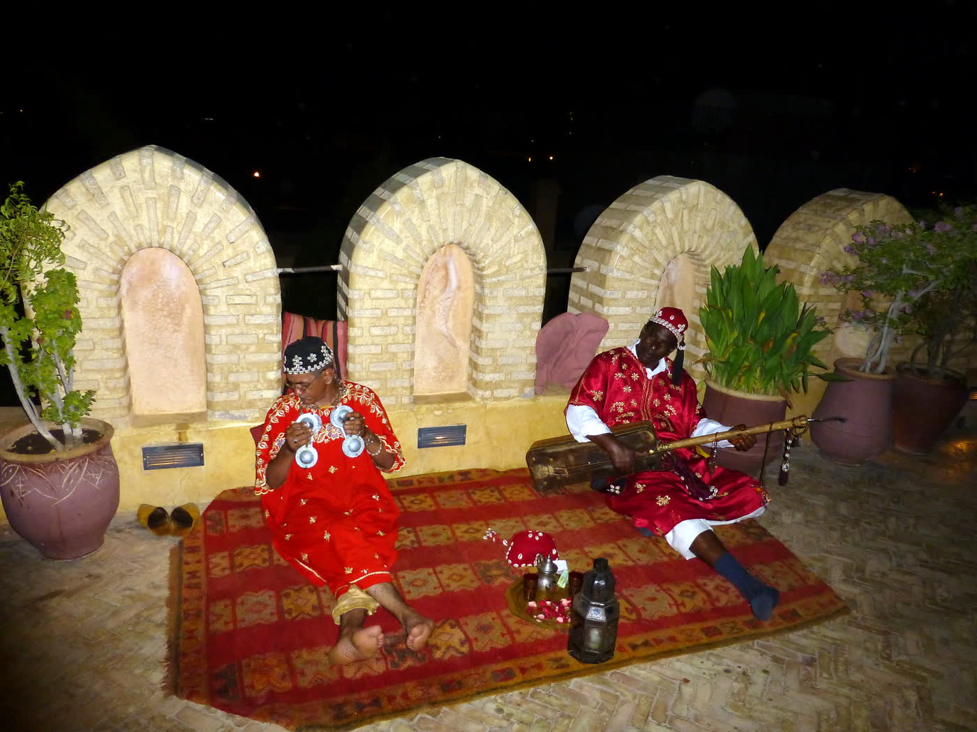 Two musicians in red embroidered robes and caps sitting on a red carpet on a rooftop terrace, one with metal castanets and one with a long-necked string instrument