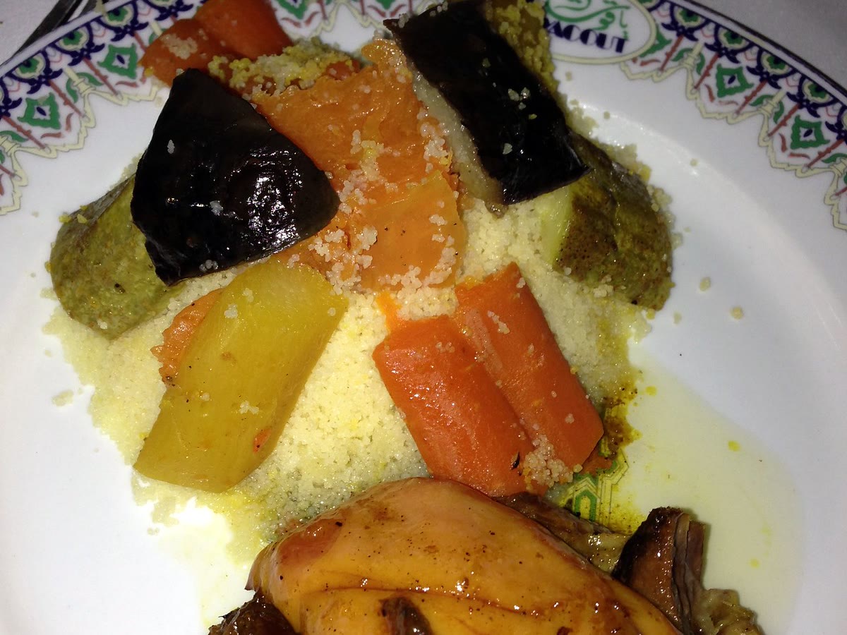 Steamed couscous topped with carrot, turnip, aubergine and courgette beside a piece of glazed meat, on a plate printed with the restaurant’s name