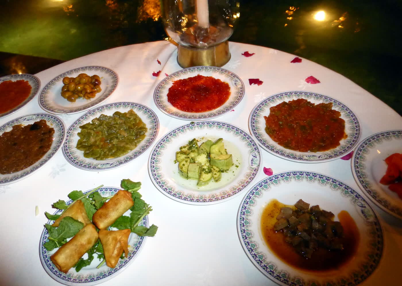 A round table with a white cloth, a candle lantern and nine small plates of Moroccan salads, olives and fried pastry rolls, on patterned porcelain