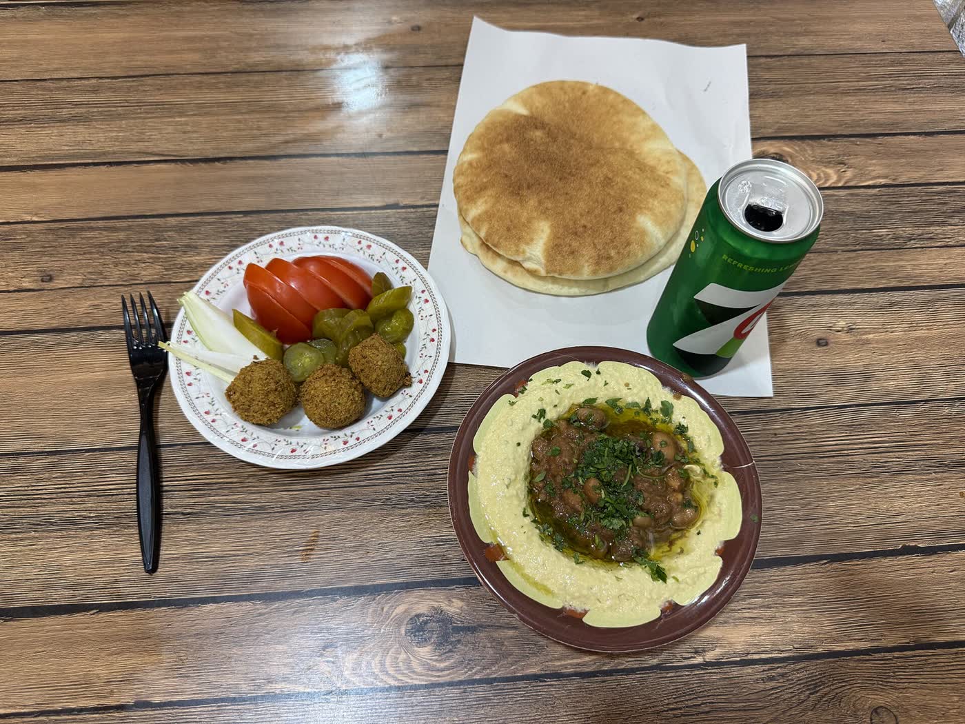 A table with a dish of hummus topped with ful, a plate of falafel with tomato, onion and pickles, and a stack of pita bread
