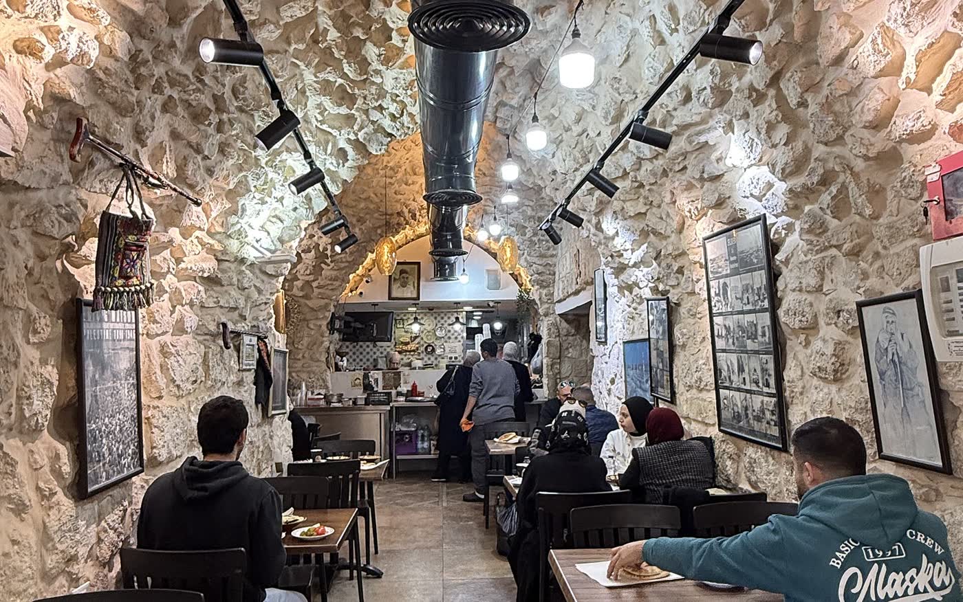 A vaulted stone dining room with framed photographs on the walls, wooden tables and diners seen from behind
