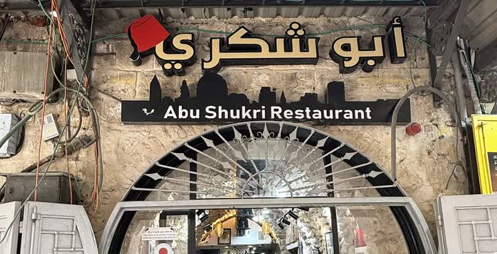 The entrance of Abu Shukri Restaurant: a black sign with the name in Arabic and English, a red fez above the first word, and a stone arch with an iron fan grille over the door
