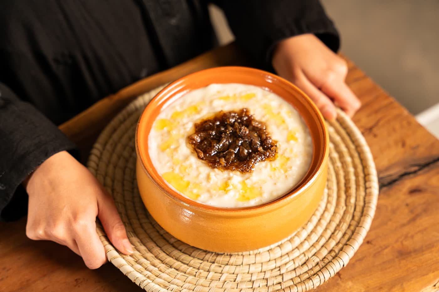 A woman’s hands hold a rope-woven mat bearing a ceramic bowl of white jareesh porridge topped with dark fried onions