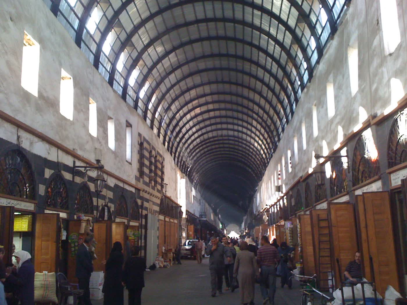 The long covered Midhat Pasha Souq in Damascus, with a curved glass-and-iron roof, shop doors on both sides and shoppers walking along the street