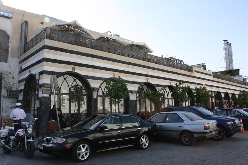 A long façade banded in black and white stone, with arched windows, small trees standing between the arches, parked cars and a policeman beside a motorcycle in front