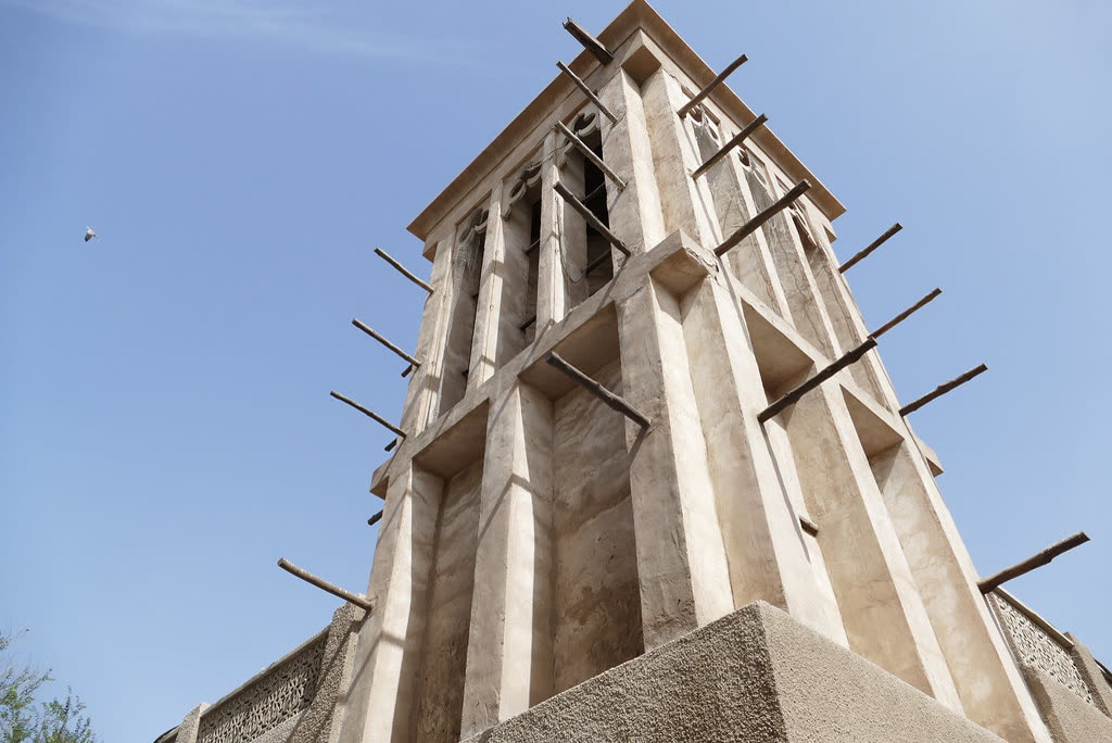 Looking up at a sand-coloured wind tower with timber beams projecting from its sides, against a blue sky
