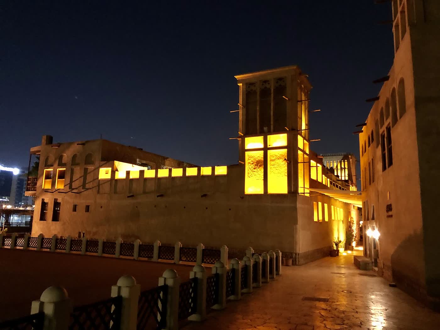 A wind tower and traditional sand-coloured houses in Al Fahidi Historical Neighbourhood, Dubai, lit in yellow against a dark night sky