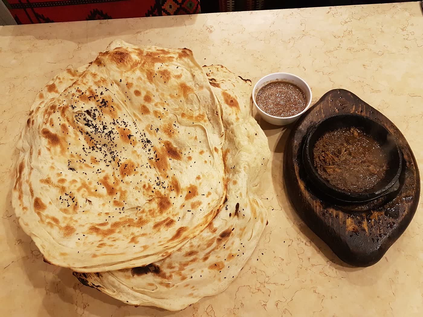 A stack of layered flatbreads sprinkled with black seeds next to a small stone pot of stew and a bowl of dip on a marble table