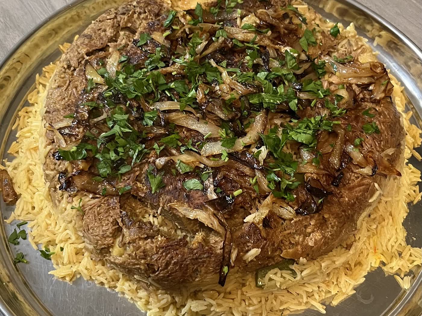 A round mound of rice topped with lamb, potato, fried onions and parsley on a metal platter