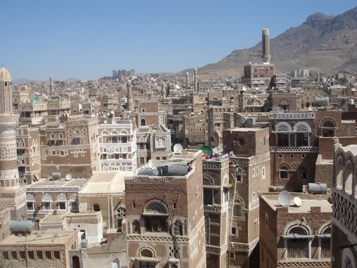 Rooftops and tall decorated brick tower houses in the Old City of Sana’a, with a minaret and mountains in the distance