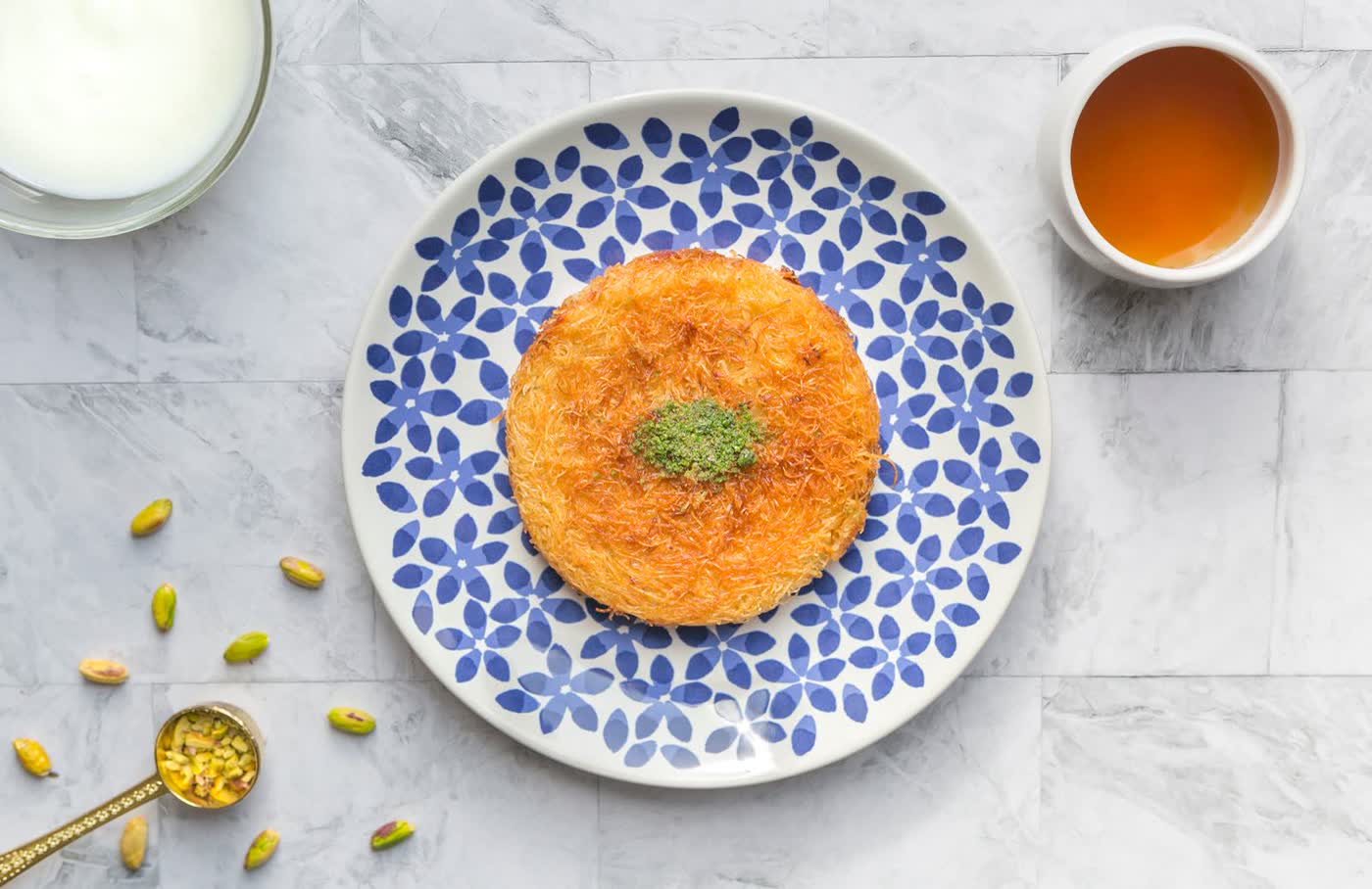 A round golden kunafa topped with pistachio on a blue floral plate, with a cup of syrup and a bowl of yoghurt beside it