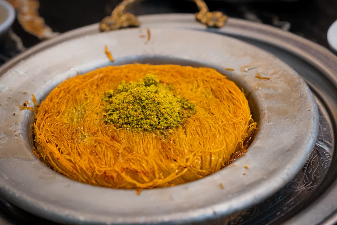 A close-up of a golden kunafa made from fine strands of pastry, topped with a mound of ground pistachio, on a metal plate