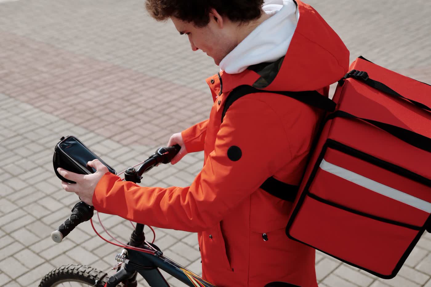 A delivery rider in an orange jacket with a large red insulated backpack checks a phone mounted on a bicycle handlebar