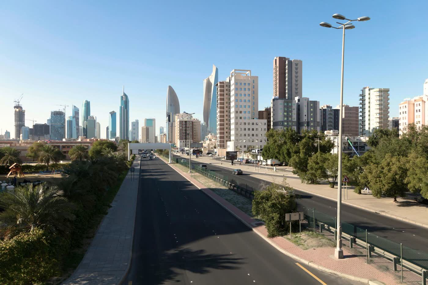 A wide road leading towards a skyline of glass towers and apartment blocks in Kuwait City under a clear blue sky