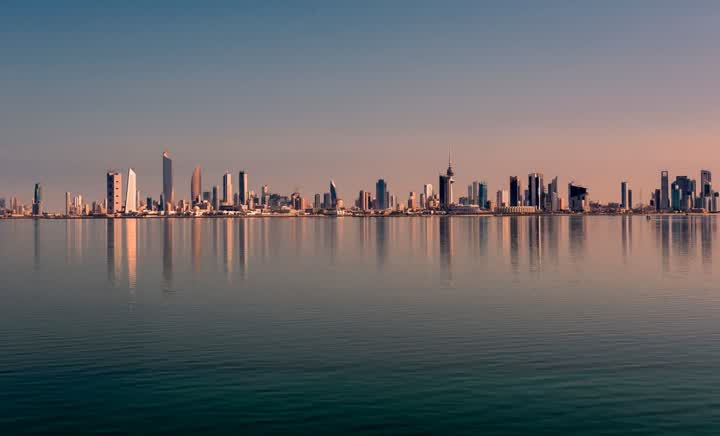 The Kuwait City skyline across calm water at dawn or dusk, reflected in the sea