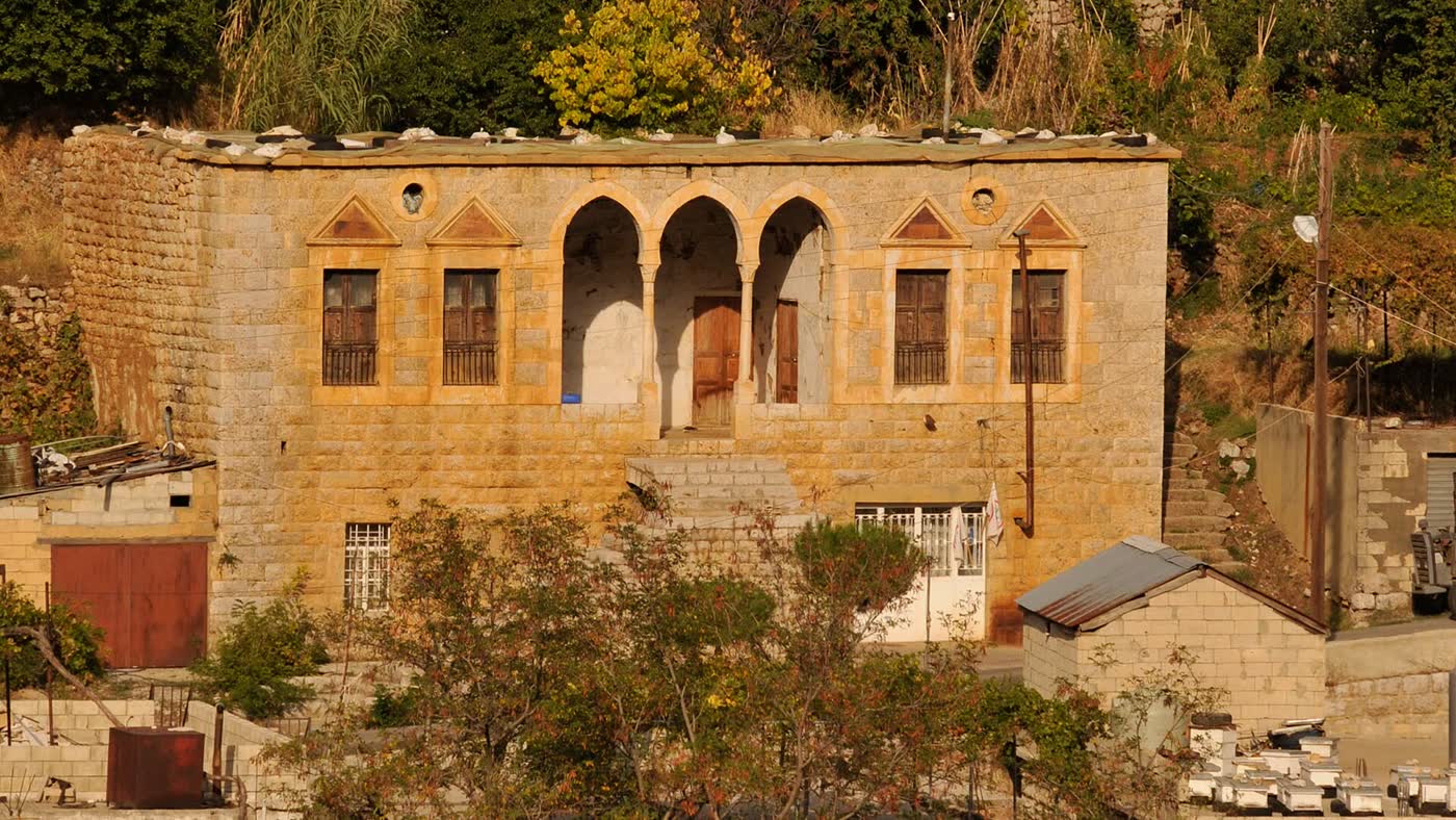 A two-storey stone house with three pointed arches across its upper floor, wooden doors and shutters, and a small terrace in front, set among trees