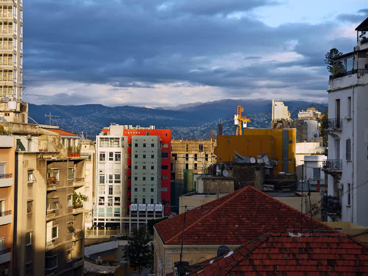 Red-tiled roofs and apartment blocks in a Beirut neighbourhood under a stormy sky, with mountains in the distance