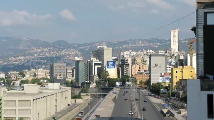 A wide Beirut road running between apartment blocks and offices, with a hazy range of hills behind